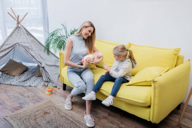 Smiling parent holding doll near daughter on couch and teepee at home 
