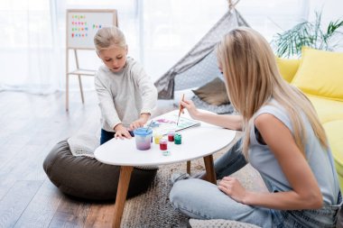 Child drawing on sketchbook near blurred mom at home 