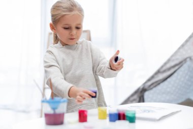 Child holding gouache near blurred sketchbook at home 