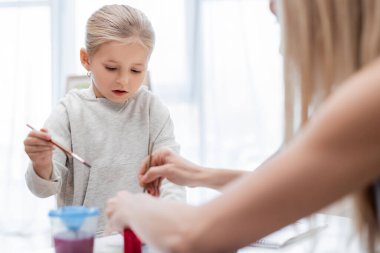 Kid holding paintbrush near blurred mom at home 