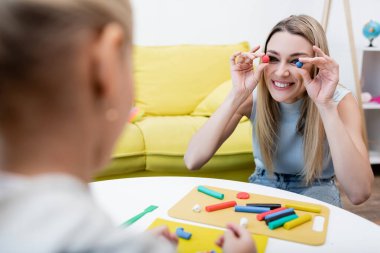 Smiling woman holding plasticine near face and blurred daughter at home 
