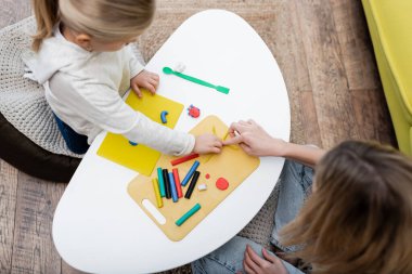 Overhead view of mom pointing at plasticine near daughter at home 