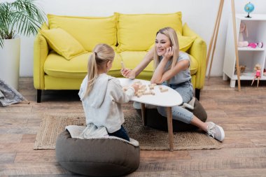 Smiling mom holding wooden block near child and coffee table at home 