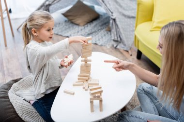 Blurred woman pointing at wood blocks game while child playing near coffee table 