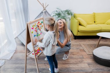 Kid fastening magnets on easel near positive mother at home 