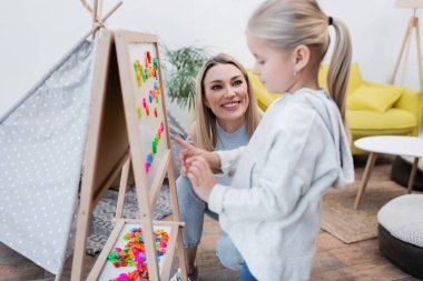 Mother looking at blurred daughter pointing at magnetic easel at home 