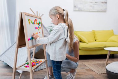Girl playing with colorful magnets on easel near mom at home 