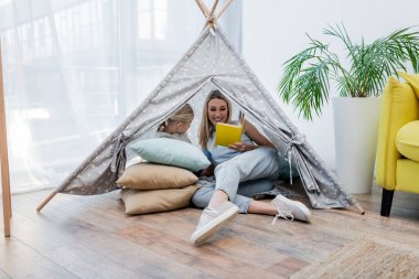 Mother reading book near child in teepee at home 