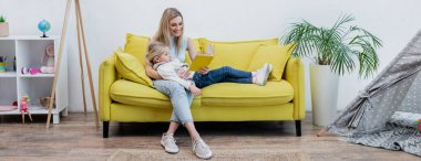 Woman reading book near kid on couch and teepee at home, banner 