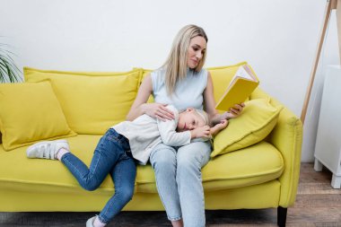 Mother reading book near kid on couch at home 