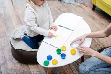 High angle view of kid pointing at sketchbook near mom and paint at home 