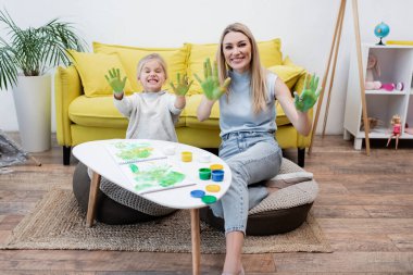 Smiling mother and child looking at camera while showing hands in paint at home 
