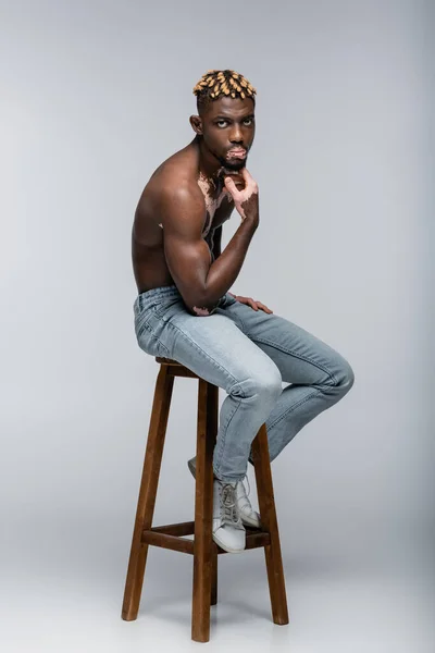 muscular african american man with vitiligo skin holding hand near chin while sitting on stool in jeans on grey