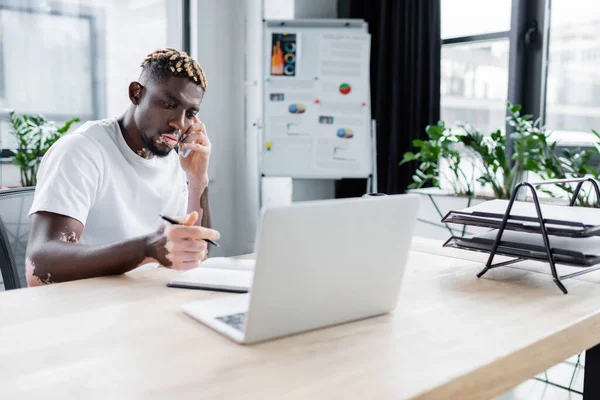 african american man with vitiligo holding pen near notebook and laptop while talking on smartphone in office