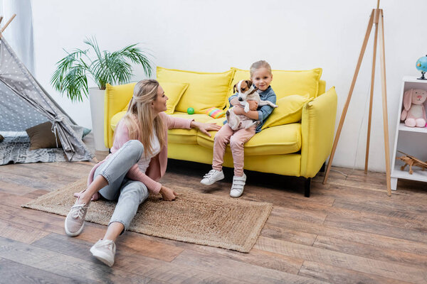 Positive kid hugging jack russell terrier near mom on floor at home 