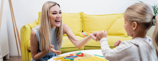 Mother pointing at plasticine near blurred daughter at home, banner 