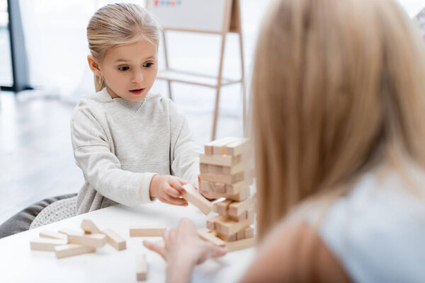 Girl holding wooden block while playing with blurred mother at home 