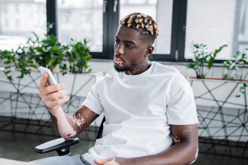 young african american man with vitiligo and trendy hairstyle looking at mobile phone while sitting in office