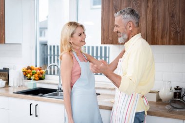 Önlüklü, gülümseyen olgun bir adam mutfakta karısıyla konuşuyor. 