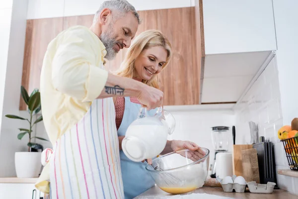 Önlüklü olumlu bir adam mutfakta yemek pişiren sarışın bir eşin yanına süt döküyor. 