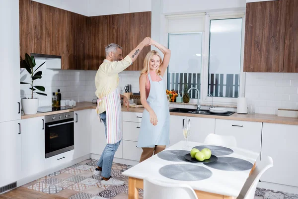 Önlüklü gülümseyen bir kadın kocayla mutfakta şampanya eşliğinde dans ediyor. 