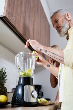 Dövmeli bir adamın mutfaktaki blender 'a meyve dökmesinin düşük açılı görüntüsü 