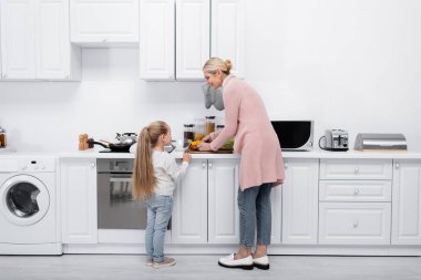 Gülümseyen olgun bir kadın ve küçük torunu modern mutfakta birlikte yemek pişiriyorlar.