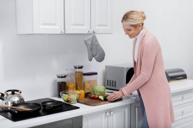 Gülümseyen ev hanımı mutfakta salata hazırlarken salatalık kesiyor.