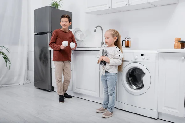 Garçon Souriant Tenant Tasses Près Soeur Avec Couverts Dans Cuisine — Photo