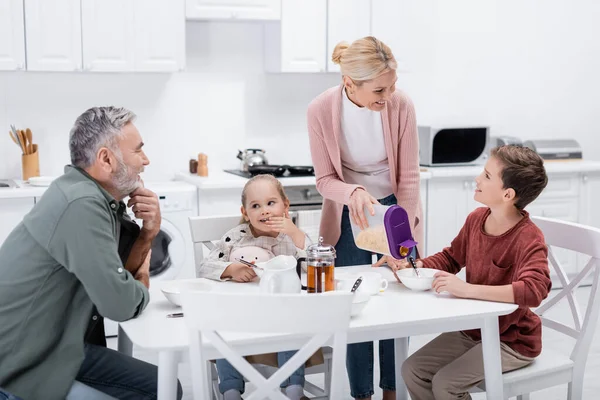 middle aged woman with corn flakes smiling near grandchildren and husband in kitchen