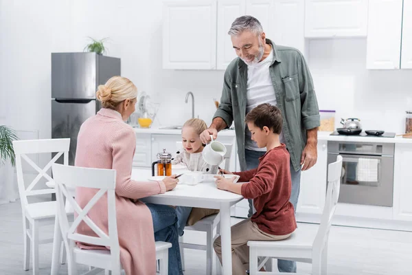 Happy Man Pouring Milk Wife Grandchildren Breakfast — Stock Photo, Image