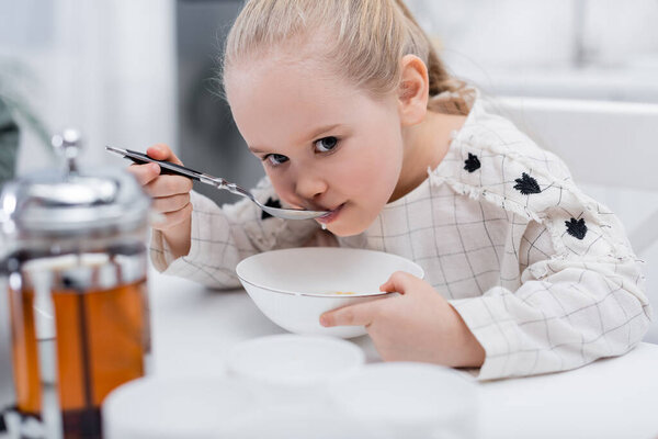 girl having breakfast in kitchen near blurred teapot and cups