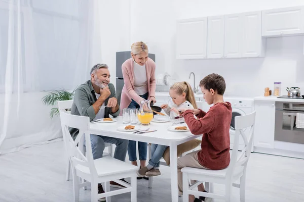 Vrolijke Vrouw Serveert Pannenkoeken Voor Kleinkinderen Echtgenoot Tijdens Het Ontbijt — Stockfoto