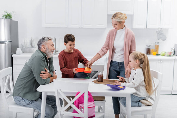 Улыбающаяся женщина с бутербродами рядом с внуками с коробками на кухне