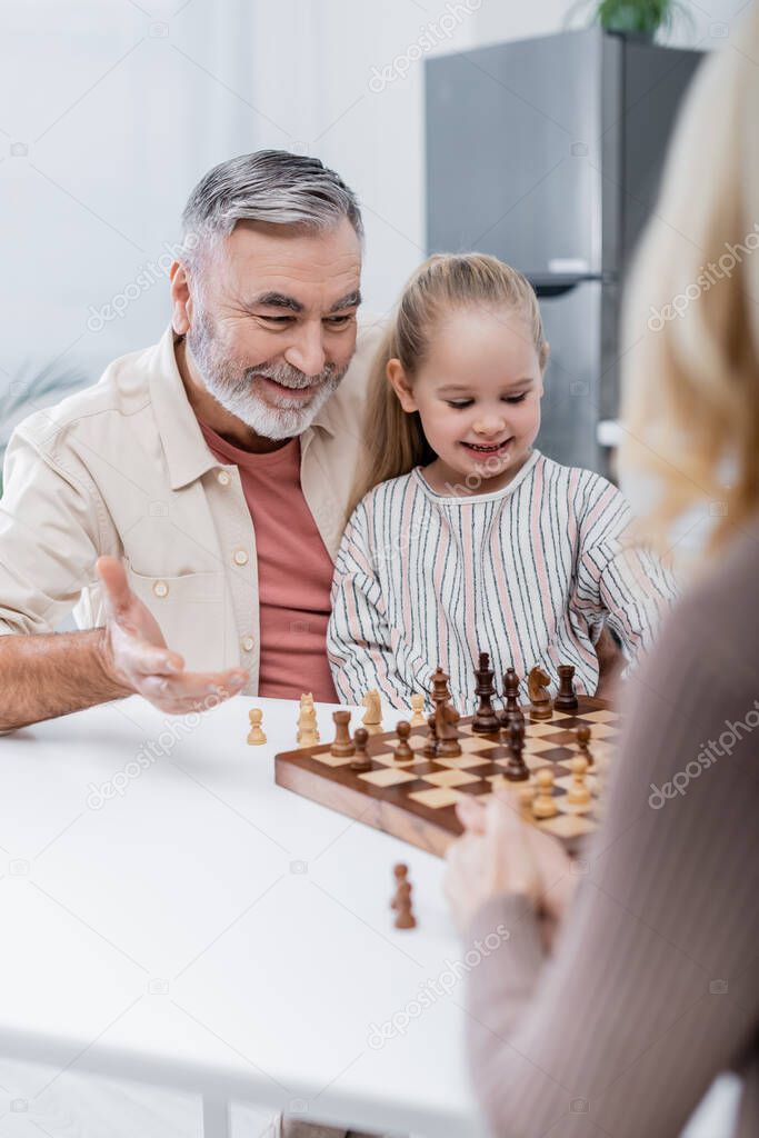 smiling senior man pointing with hand at chessboard near granddaughter and blurred wife