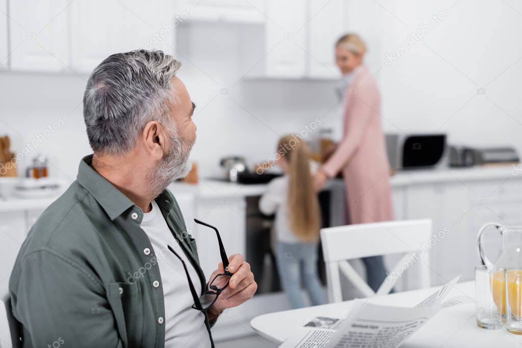 senior man with eyeglasses and newspaper looking at blurred wife and granddaughter preparing breakfast in kitchen