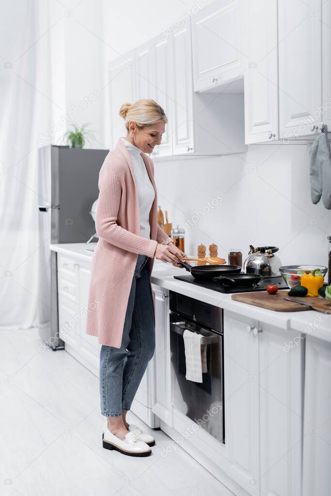 full length view of happy middle aged woman cooking pancake near fresh vegetables 