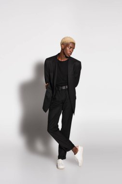 Siyah takım elbiseli ve beyaz spor ayakkabılı Afro-Amerikan bir adamın, elleri arkada, gölgeli, gri arkasında poz verişi.