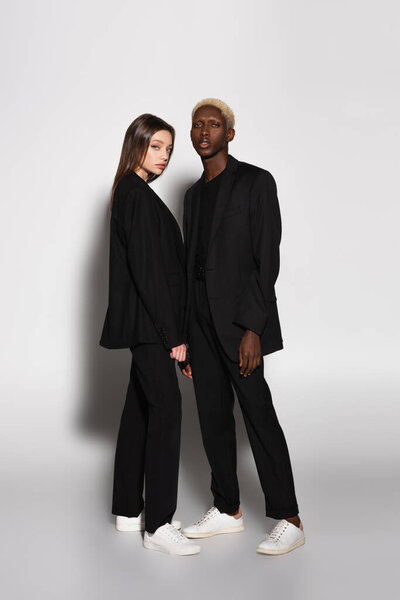 полный вид на модную межрасовую пару в черных костюмах и белых кроссовках на сером с тенью