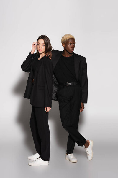 красивая и стильная женщина смотрит в камеру рядом с блондинкой африканский американец на сером с тенью