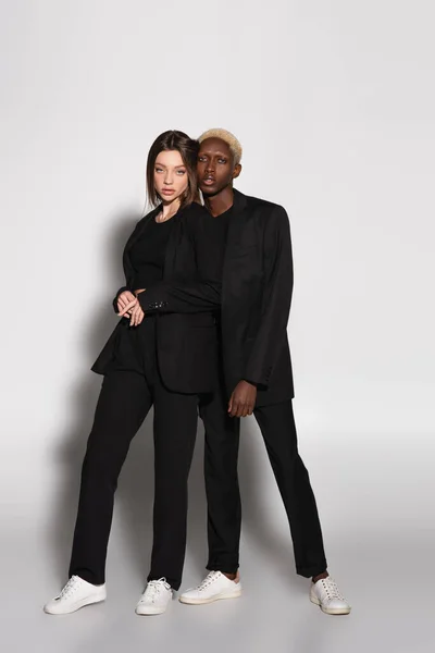 黒のスーツを着たスタイリッシュな異人種間のカップルの完全な長さのビューは 影と灰色にポーズ — ストック写真