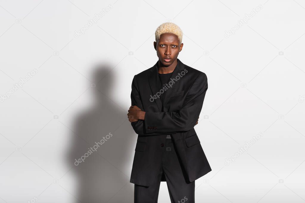 blonde african american man in black suit standing with crossed arms on grey with shadow