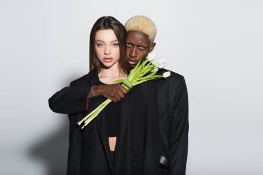 blonde african american man holding white tulips near stylish brunette woman on grey with shadow clipart