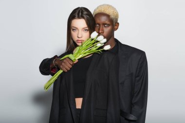 Beyaz saçlı Afrikalı bir adam yanında beyaz laleler tutan gri elbiseli, esmer bir kadın.