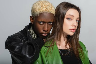 brunette woman and blonde african american man in leather jackets looking at camera isolated on grey clipart