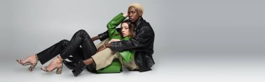 stylish brunette woman and blonde african american man looking at camera while sitting on grey, banner clipart