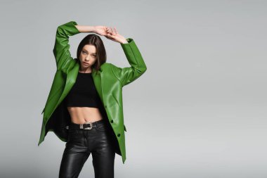 Siyah bluzlu ve yeşil deri ceketli moda bir kadın ellerini başının üstüne koyup gri üzerinde duruyor.