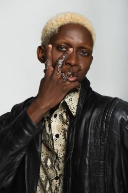 Yılan şekilli yüzükler takmış şık Afro-Amerikan bir adam. Gri renkte, yüzü kapalı poz veriyor.