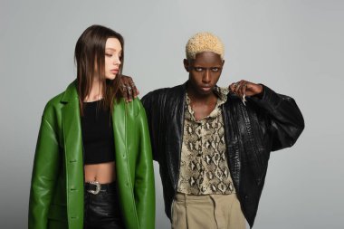 blonde african american man in black jacket and snakeskin print shirt posing near brunette woman isolated on grey clipart