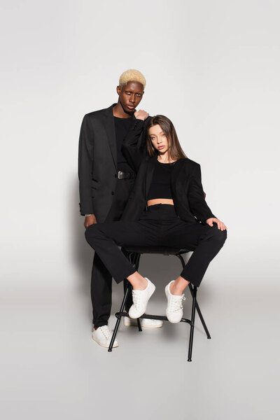 женщина в черном костюме и белых кроссовках сидит на стуле рядом с блондинистым африканским американцем на сером с тенью